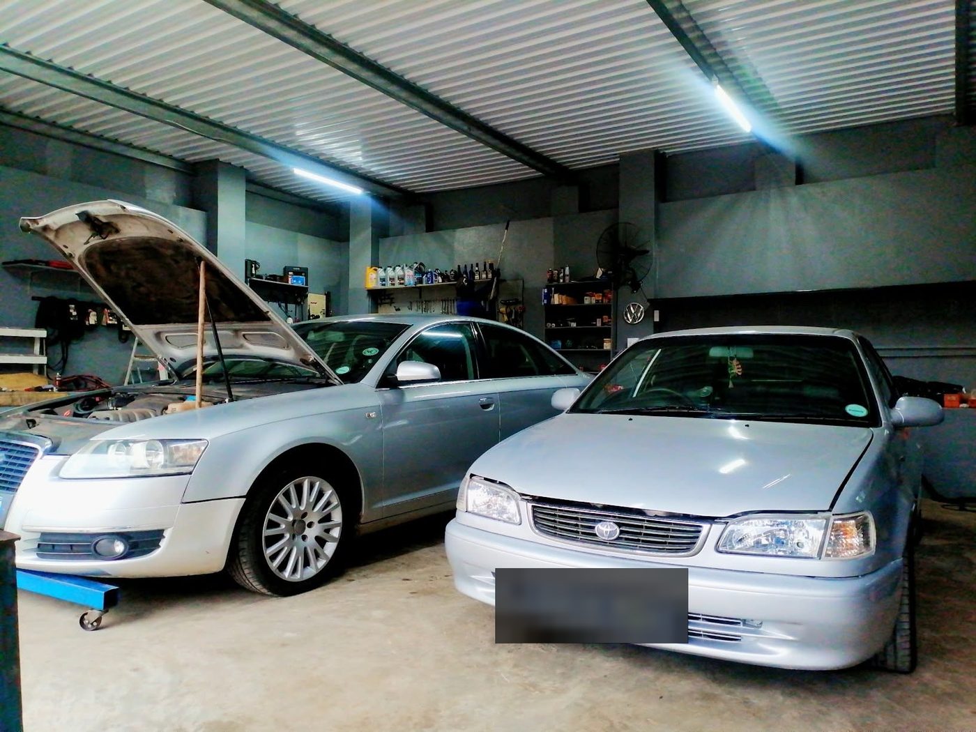Inside the workshop: a silver Audi A6 with its bonnet raised beside a silver Toyota Corolla, with a wall of tools, oils and parts on shelves behind them.